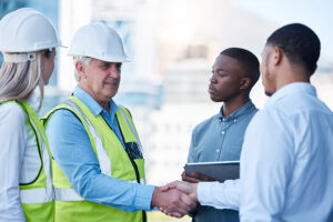 team management: Building, handshake and men in deal for architecture, planning and agreement at construction site. Project, collaboration and civil engineering team shaking hands for urban development with welcome.