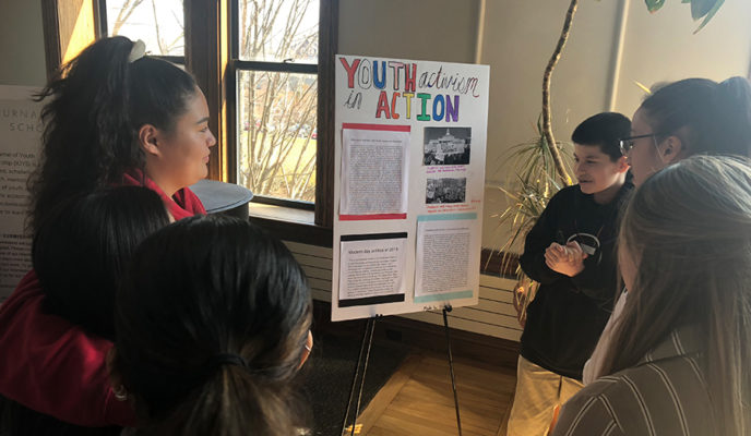 students surrounding a poster while young student gives presentation