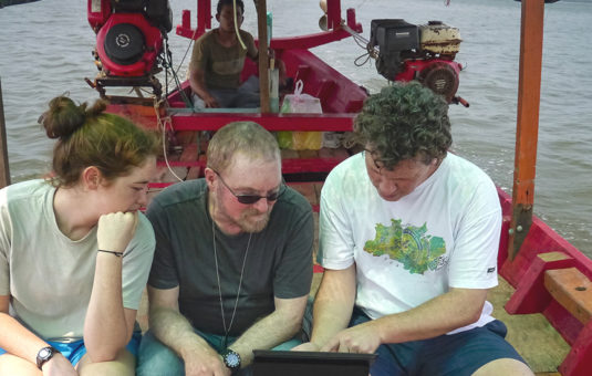 three students sitting in boat in water looking over computer