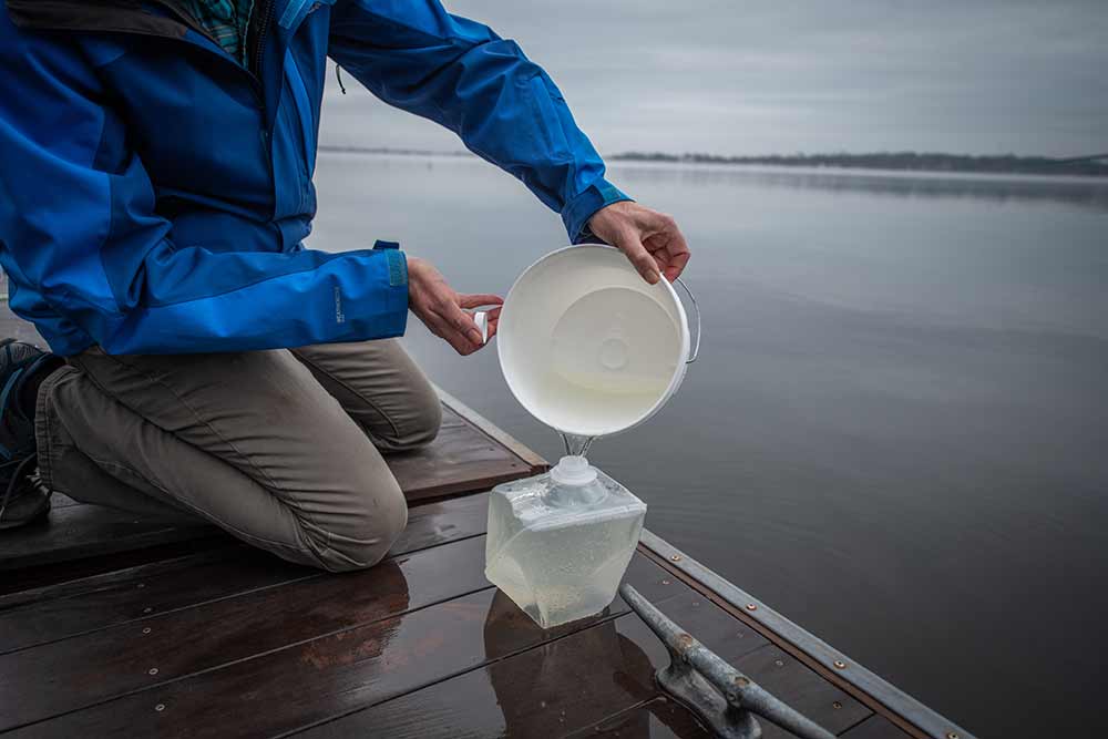 Nathan Ahlgren kneels on a dock to collects water samples to explore why viruses are killing Cyanobacteria