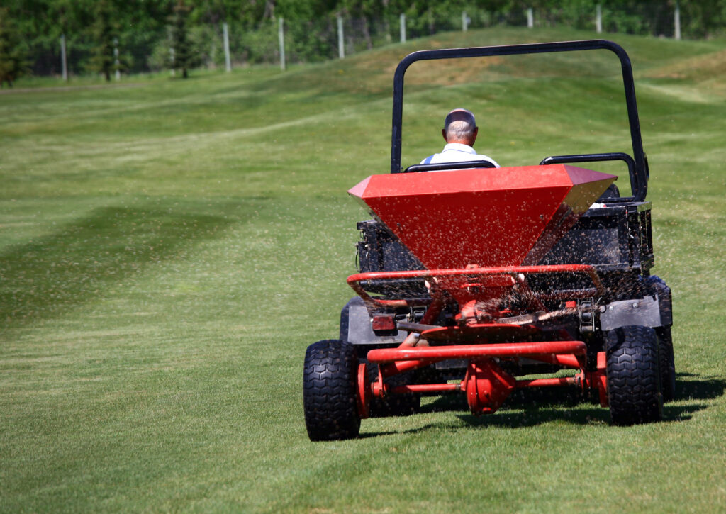 A man on a riding machine spreads fertilizer on a green lawn
