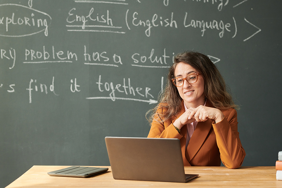 Front view portrait of adult woman as teacher looking at laptop screen in school with blackboard in background, copy space/