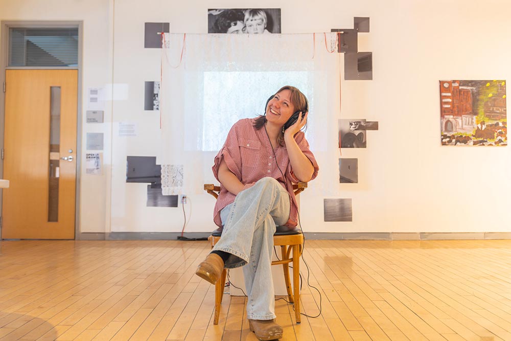 Renée Knowles seated beside the senior thesis, “POWIEDZ COŚ,” which includes lace curtains, family photos, and a projector showing a short film